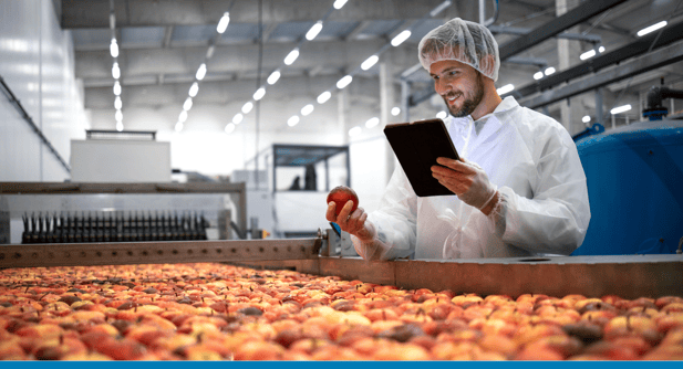 Smiling man wearing a white coat and gloves holds a tablet and examines a piece of fruit in a food manufacturing plant