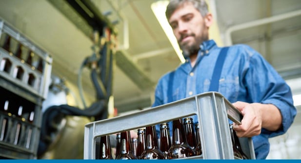 Man carries a crate of adult beverage bottles with brewing equipment in the background